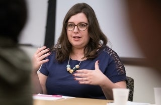 Elisabetta Ferrari speaking, while making use of her hands, to an audience. She is sitting behind a desk. She wears a navy blue blouse, glasses, and a necklace