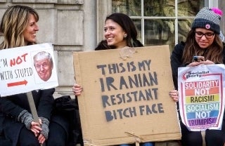 Three women seated while holding signs. The two to the left are smiling at each other, and the left-most one's white sign says " Im not with stupid", with 'not' in red, capitalised letters and has a red arrow pointed to a cut-out of Donald Trump's face. The one in the middle has a cardboard sign which reads "This is my Iranian Resistant Bitch Face". Finally, the lady to the right, who is typing on her phone, is holding a sign that reads "Solidarity not Racism! Socialism not Trumpism" on a rainbow background