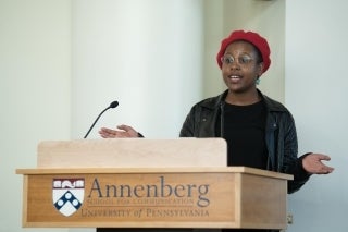 Florence Madenga speaking behind a wooden podium. The podium has a slim, long black microphone and the Annenberg School for Communication location in front. She is wearing clear glasses and a red hat.