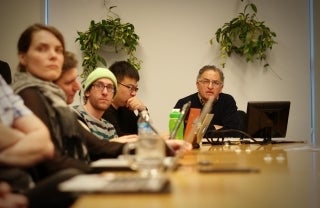 Line of students and Joseph Cappella seated at a table. They are two hanging plants on the wall behind Cappella.