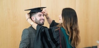 Woman in graduation regalia putting mortarboard hat onto student
