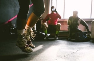 Side-view of someone's body from the knees down as they jump in a gym. They are wearing socks and sneakers. The gym is padded with black material. To the person's left are two people sitting beside each other, with one drinking water. Behind them is a big window, and we can see it is daytime. photo credit Dylan Nolte / Unsplash