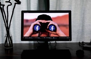 Face on computer screen looking through binoculars that have the Facebook logo on the lenses; photo credit: Glen Carrie / Unsplash