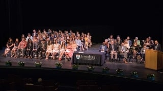 graduation 2019 1920 x 1070.jpg aerial view of the stage during graduation. A large number of students are seated in folding chairs, and faculty are on the right in folding chairs. A speaker is at the podium.