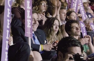 still01 1920x1250 CROP.jpg A crowd of people looking in front of them. Some are holding up long, purple signs with white text that says 'Michelle' in upper-case letters.