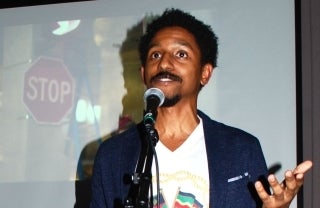 Aymar Jean Christian speaking into a microphone on a stand. Behind him is a projected image/video still of somewhere with a visible stop sign.