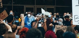 Photo Credit for Josh Hild, Unsplash. Photo is a group of people wearing masks, some holding up cardboard sign with black text, and they surround a man in the center who is talking to them with a megaphone. Some people have their fist up, and some are recording on their phones.
