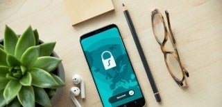 Desk with, from left to right, a succulent, pair of wireless headphones, a cellphone with a stack of sticky notes positioned above it, a pencil, and a pair of glasses lined up. The phone screen has a blue background will a simplified drawing of a part of the world in a darker blue, and on top is a white lock. At the bottom of the screen, it says 'Secured' and has a tick beside it. Photo Credit for Dan Nelson, Unsplash. 