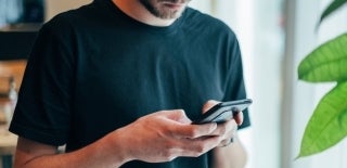 Shoulder to waist shot of a bearded person in a black shirt using their phone. They are holding up the phone with both hands and looking down at it. Photo credit for Jonas Leupe, Unsplash.