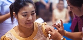 A woman in a yellow shirt sits as she is being injected in the left arm by another woman. Photo by the CDC.