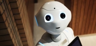 Above-angled shot of white robot with blue lights looking up at the camera. One could say the robot has a smile on its face. Photo by Alex Knight on Unsplash.