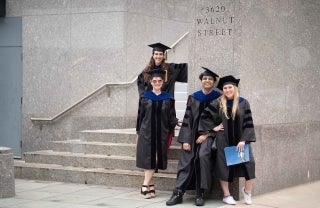 Four graduates in PhD regalia posing catalogue-style on the marble steps of 3620 Walnut St.