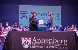 Litty Paxton handing an award to Akhil Vaidya with Annenberg logo table in front and students and faculty seated around