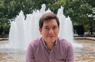 Juan Llamas-Rodriguez smiling outdoors in front of a fountain and trees behind the fountain