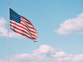 An American Flag flying on a pole against a blue sky with fluffy white clouds. Credit: Aaron Burden / Unsplash