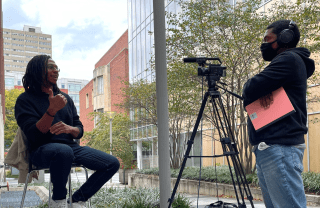 Outside, one person sits on a stool in front of a camera while being interviewed by a person standing behind a camera