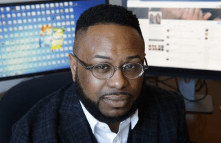 Desmond Patton sitting in front of two monitors. One monitor shows a Windows desktop, the other shows a Twitter account.