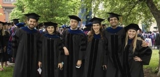 Six people posing outdoors in a line wearing PhD graduation regalia