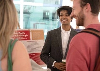 Student wearing a suit standing by a presentation board, smiling with other students