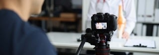 Video camera aimed at person in a white coat with a stethoscope, with another person filming