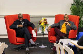 Tukufu Zuberi (left) and Brent Staples (right) sit in red chairs on a stage. Both are holding microphones and Staples is speaking into his.