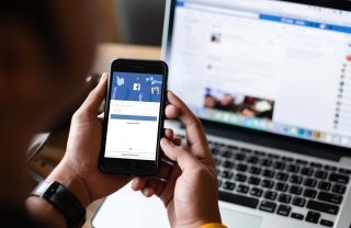 Hands of a person logging in to Facebook on a phone while a laptop with a blurred Facebook screen is in the background