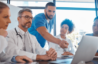 A medical team in an office sits around a table with a laptop; one man in scrubs points to the laptop screen