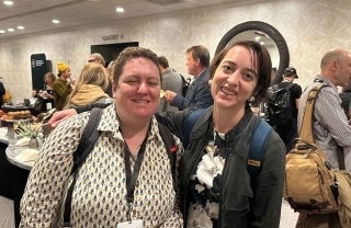 Adrienne Shaw and Jessa Lingel pose in the lobby of a hotel with various people in a crowd behind them