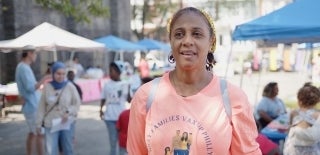 Woman in a "Vax Up Philly" shift stands in the foreground as if speaking. She is outdoors at an event with blurry people doing various things in the background.