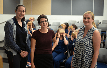 Three women standing and smiling at camera, with people in the background sitting and wearing virtual reality devices