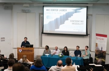 A mean speaking behind a wooden podium with the event's flyer being projected to his left. Also to his left, in front of the screen, are a table of 5 people seated. In front of them is an audience.