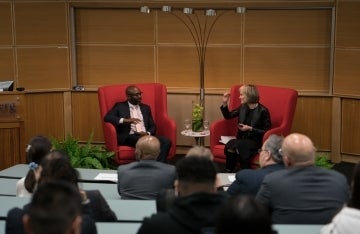 Willow Bay and John L. Jackson Jr. speaking in front of an audience. They are taboo seated in red chairs with high backs, and decor surrounds them.