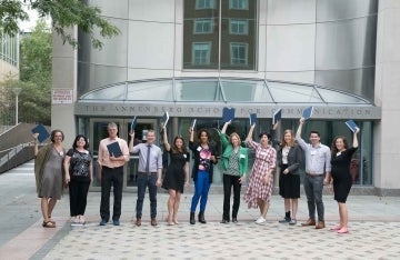 Ten people standing in a row outside the Walnut St. entrance to the Annenberg School Building, holding blue books in the air