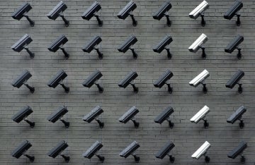 5 rows of security cameras on a brick wall, one column has white cameras and the others are black and gray 
