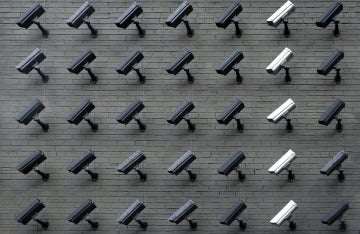 5 rows of security cameras on a brick wall, one column has white cameras and the others are black and gray