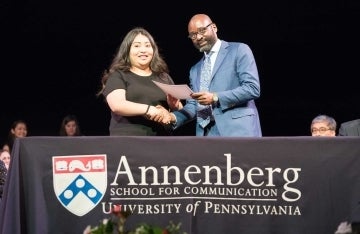 Dean John L. Jackson, Jr. shaking hands with a graduate and handing her a certificate while they both smile at the camera. They stand behind a tablecloth with the Annenberg logo on it.