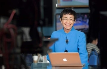 Maria Ressa sitting on a TV set with a laptop open in front of her, smiling