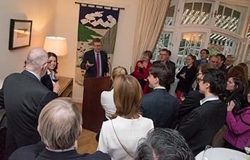 Group of people at a party indoors standing around a podium where a man speaks.