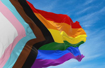 A Progress Pride Flag blows in the wind against a blue sky with several clouds