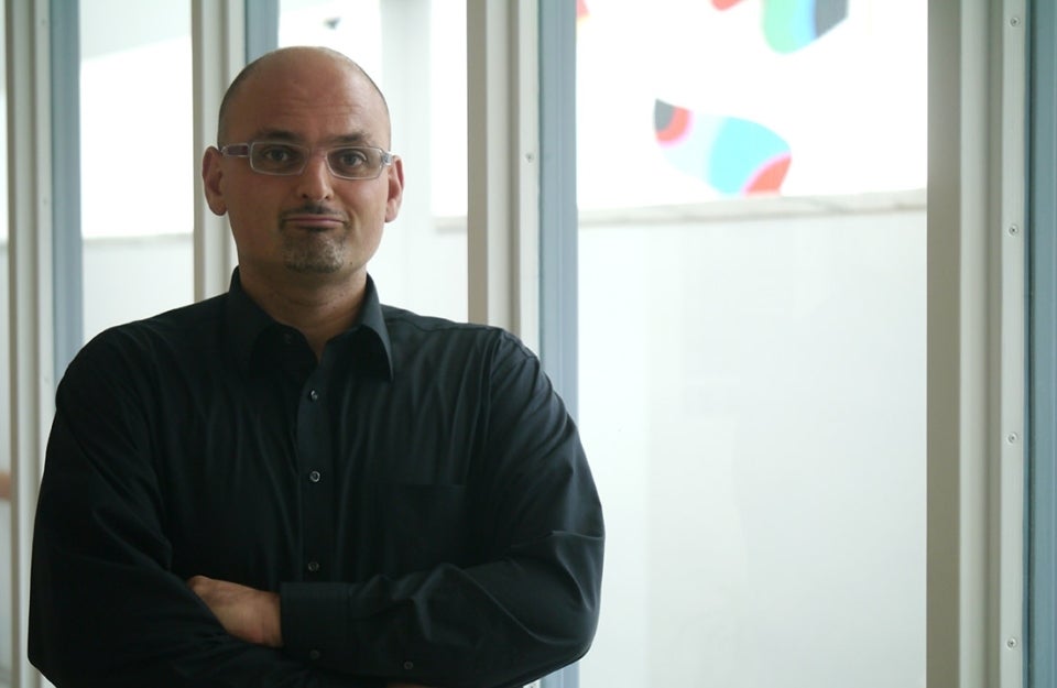 Marwan Kraidy poses in a black shirt with his arms folded in front of big glass windows.