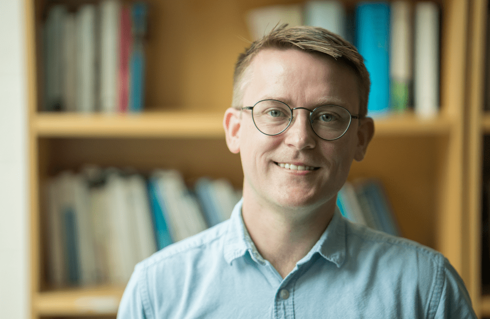 t: David Lydon-Staley, wearing round glasses and a light blue button-up shirt, smiling in front of a wooden bookshelf filled with books.