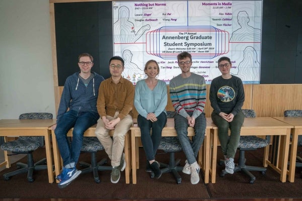 Five students seated on tables in front of a screen reading "The 7th Annual Graduate Student Symposium"