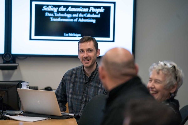 Man smiling at two other people, one of whom is laughing. The screen behind him reads "Selling the American People" 
