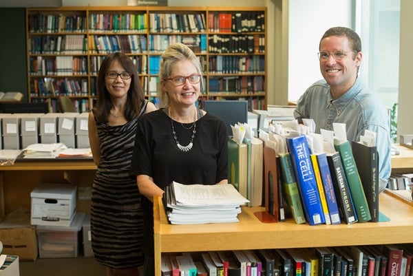 From left to right, Min Zhong, Sharon Black, and Jordan Mitchell pose behind a hip-level book shelf. Min Zhong is standing slightly behind the other two who are beside each other. They are all smiling. The background is a bookshelf-wall that is filled.  