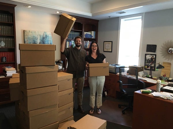 Jordan Mitchell and Laura Saywer pose with the ICA files. Saywer has a box in both her hands and Mitchell holds an empty one up with his right hand. In front of them to their right are two stacks of boxes. They are in an office setting.