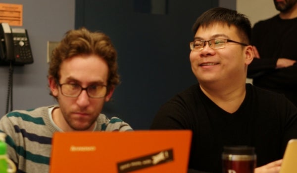 Joshua Becker and Sijia Yang seated beside each other. Becker is focused on the orange laptop opened in front of him while Yang is smiling as he looks to his right.