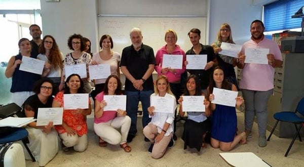 Klaus Krippendorff in the center surrounded by twenty-two European scholars who each hold a piece of paper in their hands. They are in a classroom. 