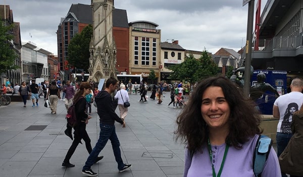 Leah Ferentinos in the lower right smiling in a square in Leicester. They are many people walking in the background, and there are many establishments.