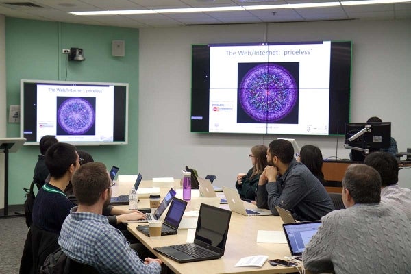 Professor Noshir Contractor in the lower right corner with his students viewing the slide deck from a class speaker. They are in a classroom with two different sized monitors to view.