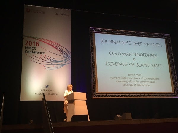 Barbie Zelizer stands on stage behind a podium. Behind her is a vertical banner for the 2016 IAMCR Conference and to her left is the projection screen. The title of the presentation is 'Journalism's Deep Memory: Cold War Mindedness & Coverage of Islamic State'  and below it is 'Barbie Zelizer, Raymond Williams Professor of Communication, Annenberg School for Communication, University of Pennsylvania'
